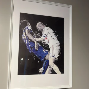 Zidane ikonisk moment tavla  - Konstverk som visar en dramatisk fotbollsscen med två spelare. Den ena bär en blå kortärmad tröja med gula detaljer, medan den andra har en vit kortärmad tröja med röda och blå inslag. Perfekt för fotbollsälskare som vill ha en ikonisk bild på väggen.