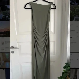 Grön långklänning med drapering - Elegant grön långklänning med draperad detalj på sidan. Klänningen har en stilren design med en axelbandslös stil som ger en sofistikerad look. Perfekt för speciella tillfällen.