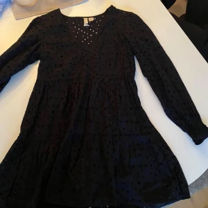 Svart klänning med hål - Snygg svart klänning med prickigt mönster och v-ringad design. Den har långa ärmar och en luftig passform som ger en skön känsla. Perfekt för en stilren look.