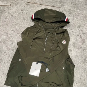 Grön vindjacka från Moncler - Säljer en snygg grön vindjacka från Moncler med huva och dragkedja. Jackan har Moncler-logga på ärmen och ribbade detaljer i rött, vitt och blått på huvan. Perfekt för blåsiga dagar! Skriv vid frågor, kvitto finns o den är utan skador. Nypris är väl ca 8 har ja för mig, ha en bra dag om du har fet röv ❤️