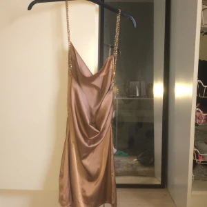 Champagnefärgad klänning från Shein - Snygg champagnefärgad klänning från Shein med kedjeaxelband. Klänningen har en draperad design och är perfekt för en festlig kväll. Materialet ger en lyxig känsla och den korta längden gör den extra chic.
