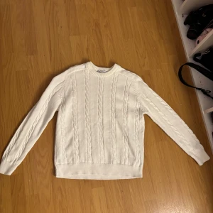 Vit stickad tröja - Säljer en stilren vit stickad tröja med kabelstickat mönster. Tröjan har långa ärmar och ribbade kanter vid ärmslut och nederkant. Perfekt för kyligare dagar!