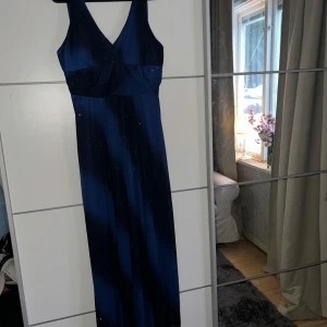 Mörkblå långklänning från Dollar - Elegant mörkblå långklänning från Dollar med v-ringning och ärmlös design. Perfekt för en kväll ute med stil. Klänningen har en subtil glittrig effekt som ger en extra touch av glamour.