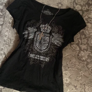 Svart t-shirt med tryck - Snygg svart t-shirt med ett detaljerat tryck av en sköld med vingar och en krona. Texten 'Sacred' finns under motivet. Perfekt för en cool och avslappnad stil.