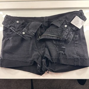 Svarta jeansshorts med slitningar - Säljer ett par svarta jeansshorts med coola slitningar och fransiga kanter. Shortsen har en dragkedja framtill och knappar i midjan. Perfekta för en avslappnad stil.