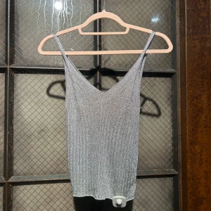 Silverfärgad glittrig linnetopp - Snygg silverfärgad linnetopp med glittrig yta och smala axelband. Perfekt för en kväll ute eller festliga tillfällen. Toppen har en v-ringad design som ger en elegant touch. Ej använd - prislapp kvar.