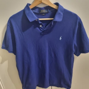 Blå pikétröja från Ralph Lauren - Pikétröja från Ralph Lauren med korta ärmar och den ikoniska loggan broderad på bröstet. Tröjan har en krage med knappar framtill.