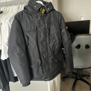 Svart parkajacka från Parajumpers - Säljer en svart parkajacka från Parajumpers med flera praktiska fickor och en snygg patch på ärmen. Jackan har en hög krage med spänne och dragkedja framtill. Perfekt för kyliga dagar!