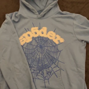 SP5DER Blå hoodie - Säljer en cool blå hoodie med en unik spindelnätsdesign och texten 'sp5der' i gult på framsidan. Perfekt för en avslappnad stil med en twist. Hoodien har en klassisk passform och är tillverkad i ett mjukt material. Storlek L passar bäst för M. Använts 2-3 gånger