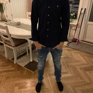 Ralph lauren skjorta - Snygg mörkblå skjorta från Ralph Lauren med klassisk krage och knappar framtill. Skjortan har en broderad logotyp på bröstet och långa ärmar. Perfekt för en stilren look.