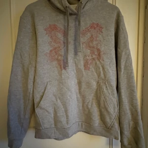 Grå hoodie med draktryck från The Cool Elephant - Snygg grå hoodie från The Cool Elephant med två röda drakar tryckta på bröstet. Tröjan har huva med snörning, känguruficka och är långärmad. Nästan aldrig använd. Pris går att diskutera.
