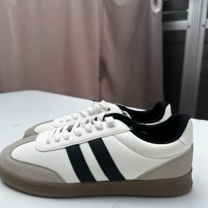 Adidas sneakers med svarta ränder - Stilrena sneakers från Adidas i vitt skinn med klassiska svarta ränder på sidorna och beige mockadetaljer vid tån. Skorna har snörning och en brun gummisula. Perfekta för dig som gillar en clean och sportig look. Helt ny