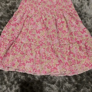 Blommig rosa volangkjol från Gina Tricot - Supersöt kort kjol från Gina Tricot i blommigt mönster med rosa, vita och gröna toner. Kjolen har smock i midjan och volangdetaljer som ger en flowy känsla. Perfekt för dig som gillar romantiska och somriga vibes!