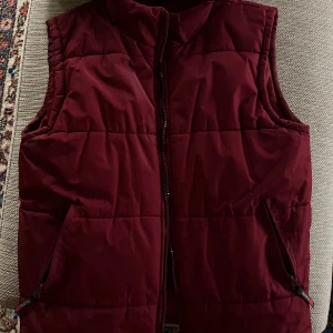 Mörkröd dunväst från Nuopai - Säljer en mörkröd dunväst från Nuopai med hög krage och två fickor med dragkedja. Västen är quiltad och har en rak passform, perfekt att slänga över en hoodie eller tröja när det är kyligt ute.