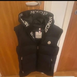 Svart dunväst från Moncler med huva - Snygg svart dunväst från Moncler med stor huva och vit logotyptext på kanten. Västen har två dragkedjefickor, quiltad design och klassisk Moncler-logga på bröstet. Perfekt för lager-på-lager och streetstyle.