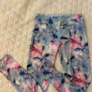 Blommiga träningsbyxor från SOC - Träningsbyxor från SOC med blommigt mönster i blått, rosa och lila. De har utskurna detaljer vid bensluten och är gjorda i ett stretchigt syntetmaterial som passar perfekt till träning.