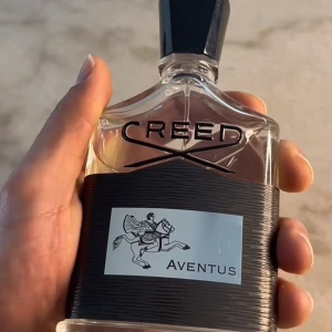 Creed Aventus parfym 100ml - Exklusiv Creed Aventus parfym i en stilren, genomskinlig glasflaska med svart etikett och svart kork. Flaskan har en elegant form och etiketten pryds av en ryttare. Aventus är känd för sin fräscha, fruktiga och maskulina doft.