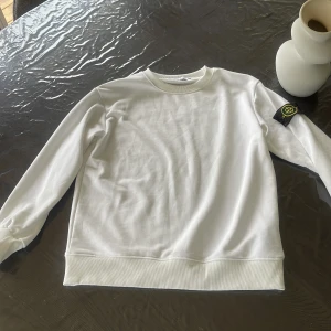 Vit Stone Island sweatshirt - Vit sweatshirt från Stone Island med klassisk rund hals och ribbade muddar. Tröjan har den ikoniska loggan på vänster ärm och är tillverkad i mjuk bomull. Perfekt för dig som gillar clean och stilren streetwear.