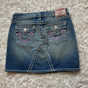True Religion jeanskjol med fickor - Snygg blå jeanskjol från True Religion med coola kontrastsömmar och  detaljer på bakfickorna. Klassisk femficksmodell med knappar och bälteshällor, perfekt för en avslappnad streetstil. Märkespatch bak i midjan och slitna detaljer för extra edge.