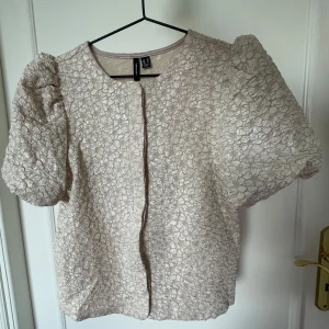 Blus med puffärmar champange/beige - Kortärmad blus i beige/chanpange från veromoda som inte finns att köpa längre. Jackan har puffiga ärmar och ett blommigt mönster i strukturen, rund halsringning och dold knäppning framtill. Perfekt för dig som vill ha något unikt och stilrent med en twist. Funkar både som bolero och blus. Passar XXS-S. Använd en gång 