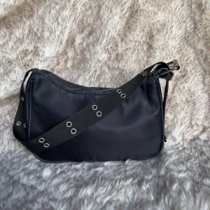 Svart axelväska med nitar och dragkedja - Cool svart axelväska i syntetmaterial med bred axelrem dekorerad med silvriga nitar. Väskan har en enkel, stilren design och stängs med dragkedja upptill. Perfekt för dig som gillar en edgy och minimalistisk look.
