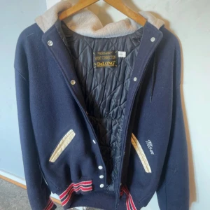 Mörkblå bomberjacka från DeLONG - Snygg mörkblå bomberjacka från DeLONG med broderad text på ryggen och kontrastfärgad huva i beige. Jackan har quiltat foder, vita detaljer vid fickorna och randiga muddar i rött och vitt. Perfekt för dig som vill sticka ut med en sportig och retro vibe.