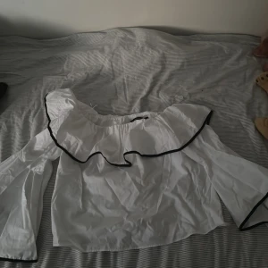 Vit blus med volang och svarta kanter - Superfin vit blus med bred volang runt axlarna och svarta kanter som ger en snygg kontrast. Blusen har vida, långa ärmar och offshoulder-modell. Perfekt för dig som vill sticka ut med en trendig och luftig topp.