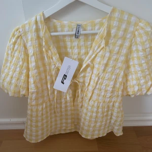 Gul och vit rutig blus  - Superfin blus i gulvitt rutigt mönster. Blusen har korta puffärmar, v-ringning och söt knytning framtill. Luftigt material som passar perfekt till sommaren