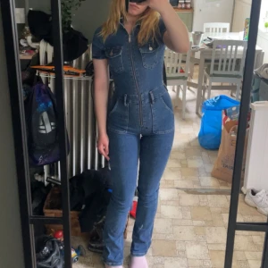 Jeans jumpsuit med dragkedja - Säljer en snygg jeans jumpsuit i blå denim med korta ärmar, markerad midja och raka ben. Jumpsuiten har dragkedja framtill, bröstfickor med knappar och klassiska jeanssömmar. Perfekt för dig som vill sticka ut med en cool och trendig look.
