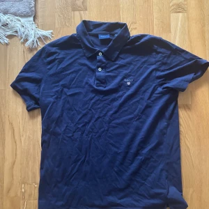 Marinblå pikétröja från GANT XL - Snygg marinblå pikétröja från GANT i storlek XL. Klassisk design med krage, två knappar och broderad GANT-logga på bröstet. Tillverkad i mjuk bomull som känns skön mot huden. Perfekt för dig som gillar stilrena och tidlösa plagg.