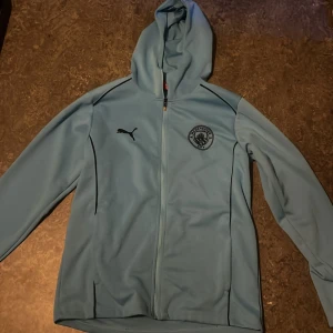 Manchester City hoodie från Puma - Ljusblå Manchester City hoodie från Puma med dragkedja och huva. Jackan har klubbens logga på bröstet, svart Puma-logga och texten 'CITYZENS' på ryggen. Perfekt för fotbollsfans som vill ha en sportig look.
