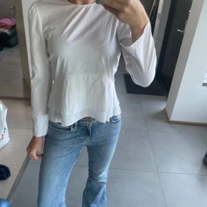 Vit volangblus från Zara Woman - Supersöt vit blus från Zara Woman med långa ärmar och volangdetaljer nertill. Blusen har en lätt utsvängd passform och är gjord i ett tunt, mjukt bomullstyg. Perfekt att styla med jeans för en clean och fräsch look.