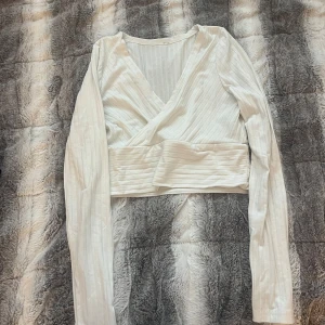 Vit omlottopp med ribbade detaljer - Säljer en vit croppad omlottopp med långa ärmar och ribbad struktur. Toppen har en djup v-ringning och bred midjeresår som ger en snygg passform. Perfekt för dig som gillar stilrena och trendiga plagg. Finns även i blå och svart!