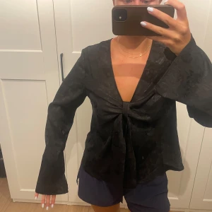 Svart omlottblus från Gina Tricot - Säljer en svart omlottblus från Gina Tricot i glansigt material med subtilt blommönster. Djup v-ringning, knyt framtill och långa vida ärmar ger en trendig look. Perfekt för dig som gillar att sticka ut med stilrena detaljer. Relativt liten i storleken 