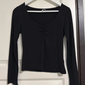 Svart långärmad topp från NLY Trend - Svart långärmad topp från NLY Trend i storlek XS. Toppen har en djup v-ringning och snygg rynkdetalj framtill som ger en cool twist. Figurnära passform och mjukt stretchigt material som känns skönt mot huden. Perfekt att styla med jeans eller kjol.