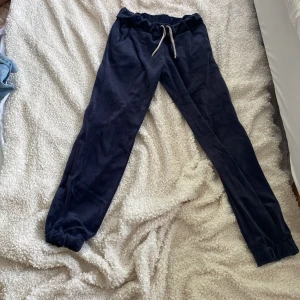 Mörkblå mjukisbyxor i fleece - Mysiga mörkblå mjukisbyxor i fleece med resår och snörning i midjan. Byxorna har muddar vid bensluten och sidofickor, perfekta för chill eller slappa dagar. Materialet är supermjukt och ger en skön känsla mot huden.