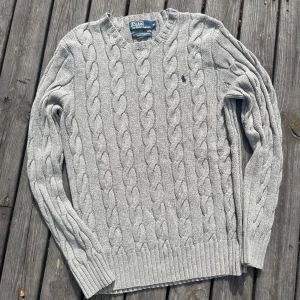 Grå handstickad cable knit Polo Ralph Lauren i Kashmir - Kabelstickad grå tröja från Polo Ralph Lauren i bomull och kashmir. Klassisk rund halsringning, ribbade muddar och diskret broderad logga på bröstet. Perfekt för dig som gillar stilren och tidlös look.