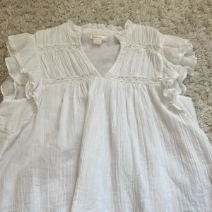 Vit blus med volang från Gina Tricot - Supersöt vit blus från Gina Tricot med volangdetaljer på ärmarna och v-ringning. Blusen har broderade spetsband och är tillverkad i ett lätt, luftigt bomullstyg. Perfekt för en fräsch och somrig look!