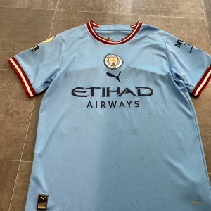 ÄKTA.Manchester City Haaland matchtröja Puma S  - Manchester Citys officiella hemmatröja från Puma med Haaland och nummer 9 på ryggen. Tröjan är ljusblå med röda och vita detaljer vid krage och ärmslut, broderat klubbmärke, Premier League-märke och 'No room for racism'-badge. Tillverkad i lätt och ventilerande polyester.