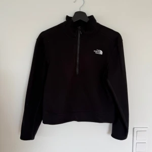 Svart fleece från The North Face - Svart fleece-tröja från The North Face med halv dragkedja och broderad logga på bröstet. Tröjan har hög krage och långa ärmar, perfekt för kyliga dagar. Enkel och stilren design som funkar till det mesta.