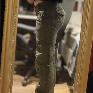Olivgröna cargopants med snörning - Säljer ett par olivgröna cargopants med flera fickor och snygga sömdetaljer. Byxorna har snörning i midjan på insidan. Passformen är loose och de har en avslappnad, baggy stil. Knappen som i midjan, ovanför dragkedjan, är lös men går att fixa om man syr till den.