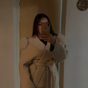 Beige kappa med äkta päls - Säljer en beige kappa med stora fluffiga fuskpälsdetaljer på axlarna och knytskärp i midjan. Jackan har en omlottmodell och bred krage som ger en lyxig vibe. Perfekt för dig som vill sticka ut med en elegant och mysig look.