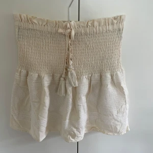 Beige kortkjol med snörning och pärlor - Supersöt beige kortkjol i fint skick med smock i midjan och volang nertill. Kjolen har snörning med tofsar och pärldetaljer, samt broderade mönster i tyget. Perfekt för sommarens alla dagar och lätt att matcha med en enkel topp.