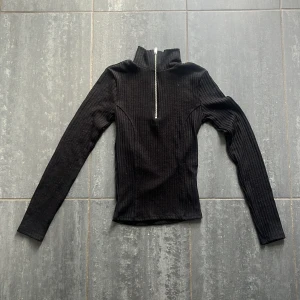 Svart ribbad polotröja från Gina Tricot - Svart ribbad polotröja från Gina Tricot med lång ärm och half zip-dragkedja framtill. Tröjan har en tight passform och hög krage, perfekt för lager-på-lager eller som statement-piece. 