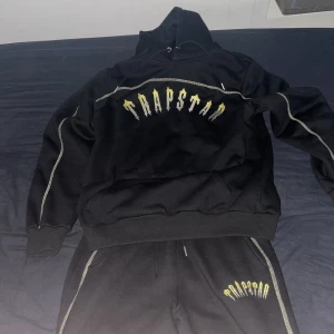 Trapstar dress Central Cee edition - Svart hoodie från Trapstar med stort gult och vitt tryck över bröstet. Hoodien har huva med snörning, kontrastsömmar längs ärmarna och en klassisk känguruficka. Perfekt för dig som gillar streetwear och vill sticka ut.