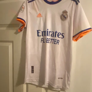 Real Madrid hemmatröja Adidas S - Säljer en officiell Real Madrid hemmatröja från Adidas i storlek S. Tröjan är vit med blå och orange detaljer på ärmslut och krage, klubbmärke på bröstet och sponsortryck framtill. Tillverkad i lätt och ventilerande material, perfekt för fotboll eller supporterliv.