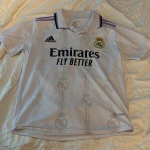 Real Madrid Vinicius Jr. 20 Adidas tröja - PRISET ÄR INTE FASTHUGGET I STEN!! Vit Real Madrid matchtröja från Adidas med Vinicius Jr. och nummer 20 på ryggen. Klassisk krage med svarta och lila detaljer, broderat klubbmärke och Adidas-logga. Tillverkad i lätt och ventilerande Climacool-material, perfekt för fotbollsplanen.