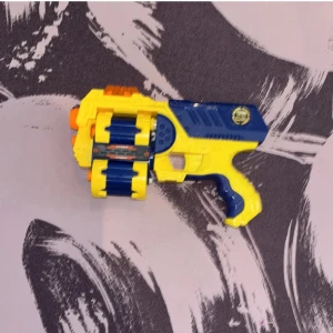 Nerf gun (leksakspistol) - Bilderna visar en leksakspistol i plast, troligen en Nerf-liknande blaster i blått, gult och orange. Den har en roterande trumma för flera skott och ser ut att vara i gott skick. Perfekt för actionfyllda lekar och tävlingar med vänner. Inga böcker syns på bilderna.