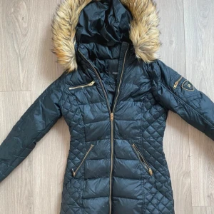 Svart Rockandblue dunjacka med fuskpäls - Snygg svart dunjacka från Rockandblue med quiltad design, guldfärgade dragkedjor och huva med fuskpäls. Jackan har flera fickor med dragkedja och Rockandblue-logga på ärmen. Perfekt för kalla dagar när du vill vara både varm och stilren.