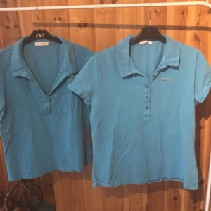 Blå pikétoppar från Lacoste - Två klassiska blå pikétoppar från Lacoste med kort ärm och krage. Toppens design har knappar framtill och den ikoniska krokodilloggan broderad på bröstet. Perfekt för en sportig och stilren look. Säljes tillsammans eller separat!